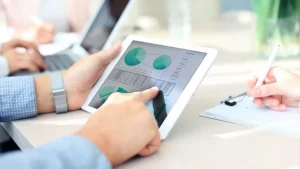 A team of professionals analyzing data on a tablet device during a meeting, with charts and graphs displayed on the screen.