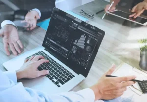 A team of professionals collaborating around a laptop screen displaying data visualizations, charts, and graphs.
